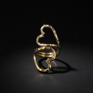 May include: A gold-colored ring with a hammered texture, featuring two heart-shaped designs. The ring wraps around the finger with an open design, creating a unique and stylish look. The ring is set against a dark background.