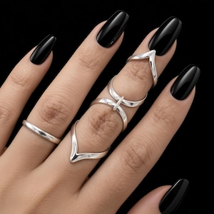 Puede incluir: Un conjunto de anillos plateados en los dedos. Un anillo es una banda sencilla, mientras que el otro es una pieza de varias partes con una punta en forma de V, una sección central circular y una base en V, unidas por una barra vertical.