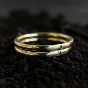 Op de afbeelding: Twee goudkleurige ringen op elkaar gestapeld. De bovenste ring heeft de letter "A" gegraveerd, en de onderste ring heeft de letter "M" gegraveerd. De ringen staan tegen een donkere, getextureerde achtergrond.