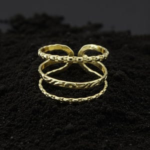 Könnte beinhalten: Ein goldfarbener Ring mit drei übereinanderliegenden Bändern. Das obere Band hat ein strukturiertes Design, das mittlere Band ein eingraviertes Muster und das untere Band ein gepunktetes Design. Der Ring ist vor einem dunklen Hintergrund platziert.