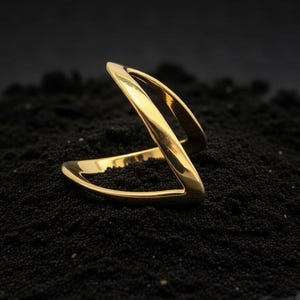 May include: A gold-colored ring with a unique, open design. The ring features a double-banded structure that curves and intersects, creating a modern and geometric aesthetic. The ring is displayed on a bed of dark, textured material, highlighting its polished finish.