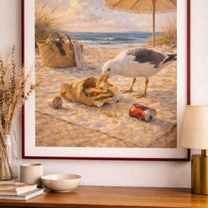 Könnte beinhalten: Ein gerahmtes Strandbild zeigt eine Möwe, die ein Sandwich an einem Sandstrand isst. Eine rote Dose, ein Weidenkorb und ein Sonnenschirm sind ebenfalls zu sehen. Das Kunstwerk ist in einem dunkelroten Rahmen gerahmt.