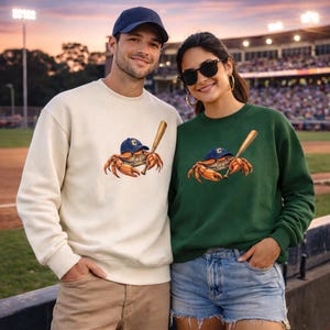 Könnte beinhalten: Cremefarbene und dunkelgrüne Sweatshirts mit einem Krabben-Design, das eine Baseballkappe trägt und einen Schläger hält. Die Krabben-Grafik ist orange, braun und blau. Die Sweatshirts werden von zwei Personen vor einem Baseballstadion getragen.