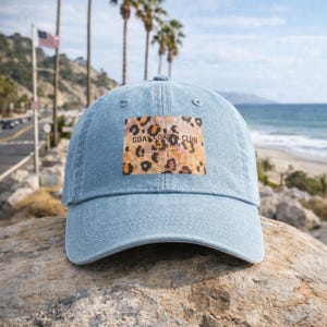 Könnte beinhalten: Hellblaue Jeans-Baseballkappe mit einem Leopardenmuster-Patch mit der Aufschrift "SOAK COAST CLUB MALIBU". Die Kappe liegt auf einem Felsen, mit Strand und Palmen im Hintergrund.