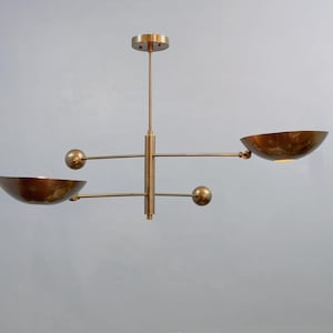 May include: A brass chandelier featuring a central vertical rod with two horizontal arms extending in opposite directions. Each arm holds a bowl-shaped shade on one end and a spherical weight on the other, creating a balanced, mobile-like structure.