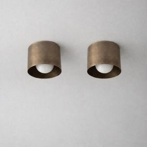 May include: Two cylindrical ceiling light fixtures featuring a brushed bronze metal finish. Each fixture houses a single, centered, spherical white glass bulb that emits a soft glow from within the deep, circular metal housing structure.