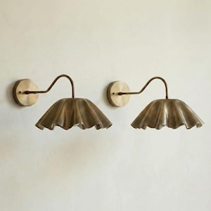 May include: A pair of wall-mounted light fixtures featuring a brushed brass finish. Each lamp has a circular wall plate, a curved arm, and a wide, ruffled, bell-shaped shade that directs light downward, creating a distinct scalloped edge profile.