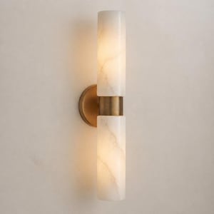Könnte beinhalten: Moderne Wandleuchte mit zwei zylindrischen Lampenschirmen aus cremefarbenem Alabaster. Die Schirme sind durch ein messingfarbenes Band verbunden und an einer kreisförmigen Messingbasis befestigt. Die Leuchte strahlt ein warmes Licht aus, ideal für die Innenbeleuchtung.