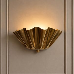 May include: A gold-toned wall sconce with a wide, scalloped, shell-like design. The fixture features a polished metallic finish and a small spherical accent at the bottom centre. It emits a warm, soft glow from the top, casting light upwards.