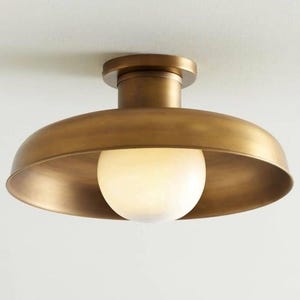 May include: A flush mount ceiling light fitting featuring a wide, circular, brushed brass metal shade with a smooth, matte finish. A single, spherical, frosted white glass globe bulb is centred within the shade, providing soft illumination.