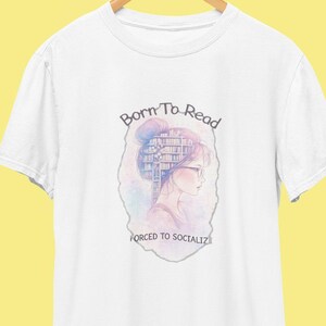 May include: White t-shirt with a watercolor illustration of a woman with a bun and glasses, with a bookshelf in her hair. The text reads "Born To Read, Forced To Socialize."