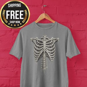 Pode incluir: Uma camiseta cinza com uma ilustração detalhada de uma caixa torácica humana. O design esquelético está centrado na camiseta, com a coluna vertebral estendendo-se para baixo. A camiseta está pendurada em um cabide de madeira contra uma parede de tijolos vermelhos.