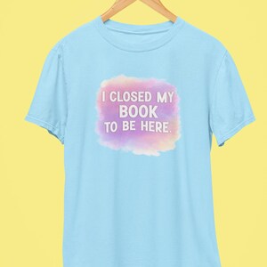 May include: Light blue t-shirt with the text "I CLOSED MY BOOK TO BE HERE" in white letters on a purple and pink cloud-like shape. The shirt is hanging on a wooden hanger against a yellow background.