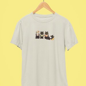 May include: Cream-colored t-shirt featuring a graphic of four black cats. Three cats are upright, reading books, while the fourth cat is lying down. The design is printed on a light-colored tee, hanging on a wooden hanger.