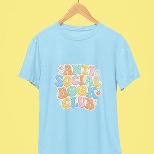 May include: Light blue t-shirt with the words "ANTI SOCIAL BOOK CLUB" in colorful, retro-style lettering. The design includes flowers and a smiley face. The shirt is hanging on a wooden hanger against a yellow background.