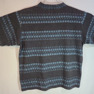 May include: A short-sleeved, dark gray t-shirt with a mock neck. The shirt features a repeating pattern of horizontal stripes and geometric shapes in light blue. The sleeves also have a similar design.