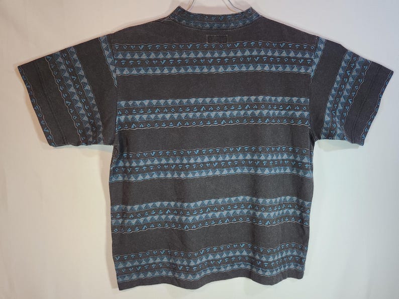 May include: A short-sleeved, dark gray t-shirt with a mock neck. The shirt features a repeating pattern of horizontal stripes and geometric shapes in light blue. The sleeves also have a similar design.