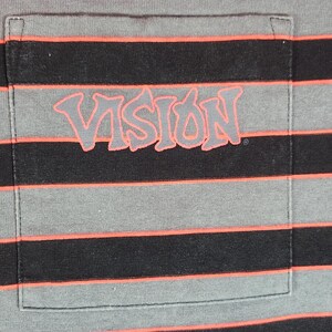 May include: Gray and black striped t-shirt with a pocket. The pocket features the word "VISION" in a stylized, coral-colored font. The shirt has horizontal stripes in gray, black, and coral.
