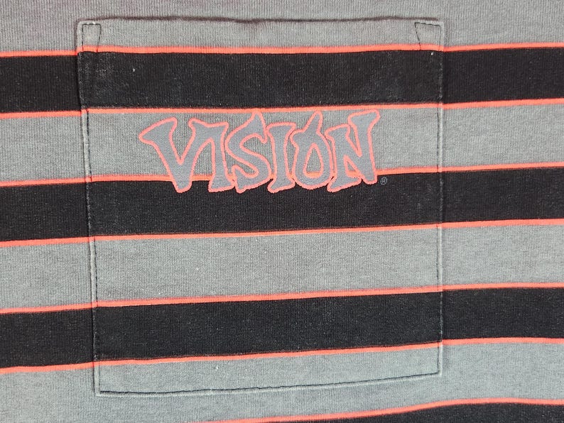 May include: Gray and black striped t-shirt with a pocket. The pocket features the word "VISION" in a stylized, coral-colored font. The shirt has horizontal stripes in gray, black, and coral.
