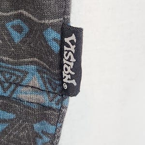 May include: Close-up of a dark gray fabric with a geometric pattern in shades of blue and gray. A black tag with the word "VISION" in white lettering is attached to the fabric.