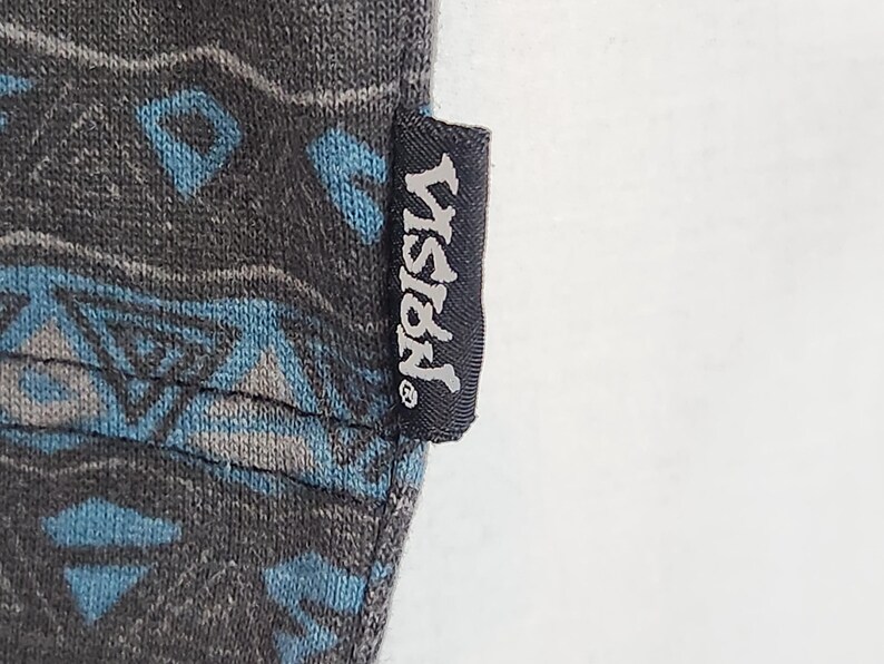 May include: Close-up of a dark gray fabric with a geometric pattern in shades of blue and gray. A black tag with the word "VISION" in white lettering is attached to the fabric.