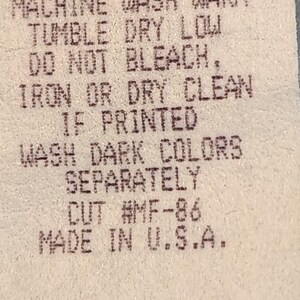 May include: Close-up of a fabric tag with printed care instructions. The tag reads "100% COTTON, MACHINE WASH WARM, TUMBLE DRY LOW, DO NOT BLEACH, IRON OR DRY CLEAN, IF PRINTED, WASH DARK COLORS SEPARATELY, CUT #MF-86, MADE IN U.S.A."