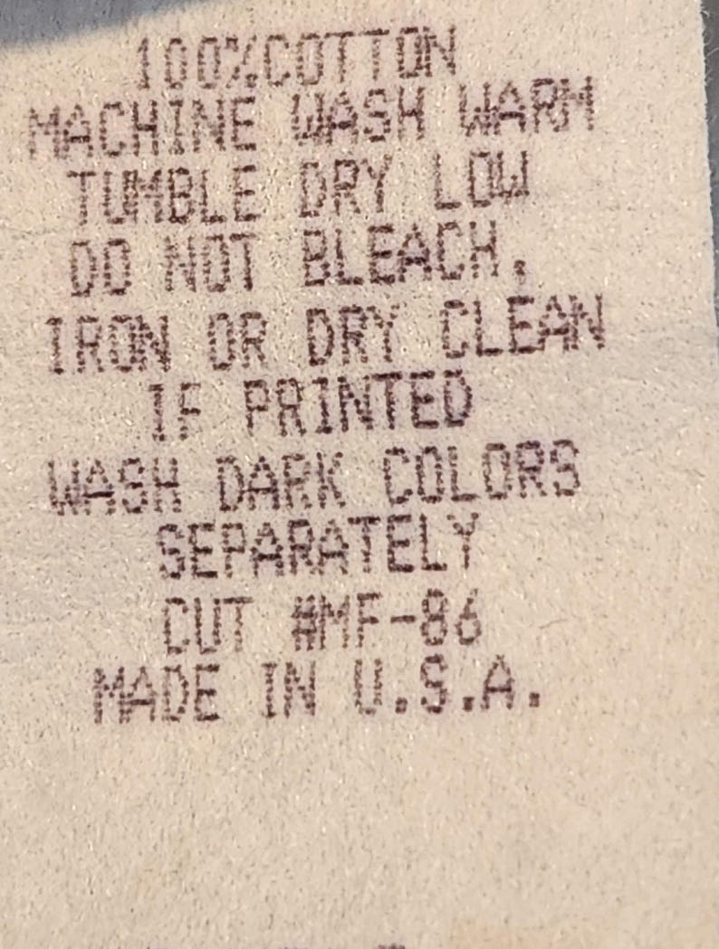 May include: Close-up of a fabric tag with printed care instructions. The tag reads "100% COTTON, MACHINE WASH WARM, TUMBLE DRY LOW, DO NOT BLEACH, IRON OR DRY CLEAN, IF PRINTED, WASH DARK COLORS SEPARATELY, CUT #MF-86, MADE IN U.S.A."