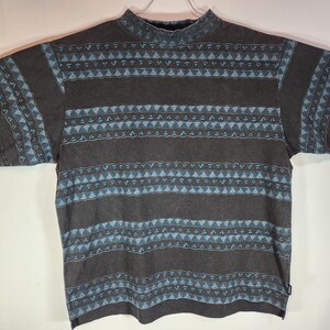May include: A gray short-sleeved shirt with a mock turtleneck collar. The shirt features a repeating pattern of horizontal stripes with geometric shapes in shades of blue and brown. The shirt is hanging on a white hanger.