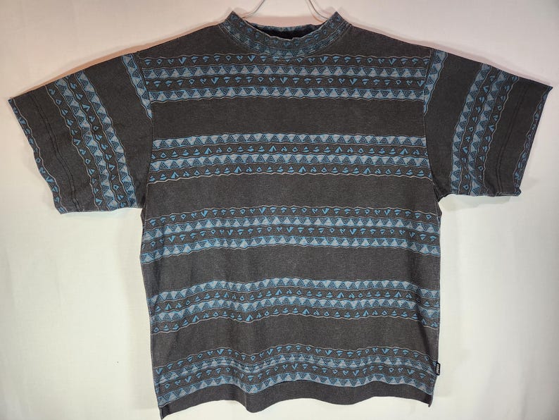 May include: A gray short-sleeved shirt with a mock turtleneck collar. The shirt features a repeating pattern of horizontal stripes with geometric shapes in shades of blue and brown. The shirt is hanging on a white hanger.