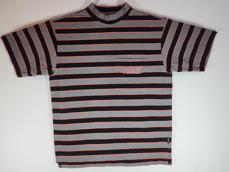 May include: A short-sleeved, striped t-shirt with a mock neck. The shirt features horizontal black and gray stripes with thin red lines. A pocket on the chest displays the word "VISION" in a stylized font. The shirt is laid flat.