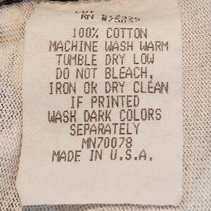 May include: A white clothing tag with black text. The tag reads "100% Cotton, Machine Wash Warm, Tumble Dry Low, Do Not Bleach, Iron or Dry Clean, If Printed, Wash Dark Colors Separately, Made in U.S.A."