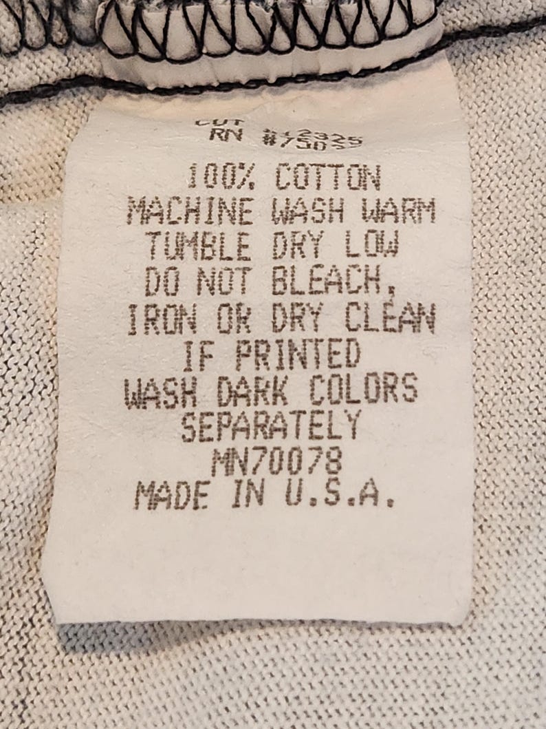 May include: A white clothing tag with black text. The tag reads "100% Cotton, Machine Wash Warm, Tumble Dry Low, Do Not Bleach, Iron or Dry Clean, If Printed, Wash Dark Colors Separately, Made in U.S.A."