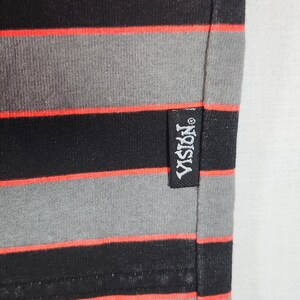 May include: Close-up of a black, gray, and red striped fabric. The fabric has horizontal stripes in black, gray, and red. A black tag with the white text "VISION" is sewn on the fabric.