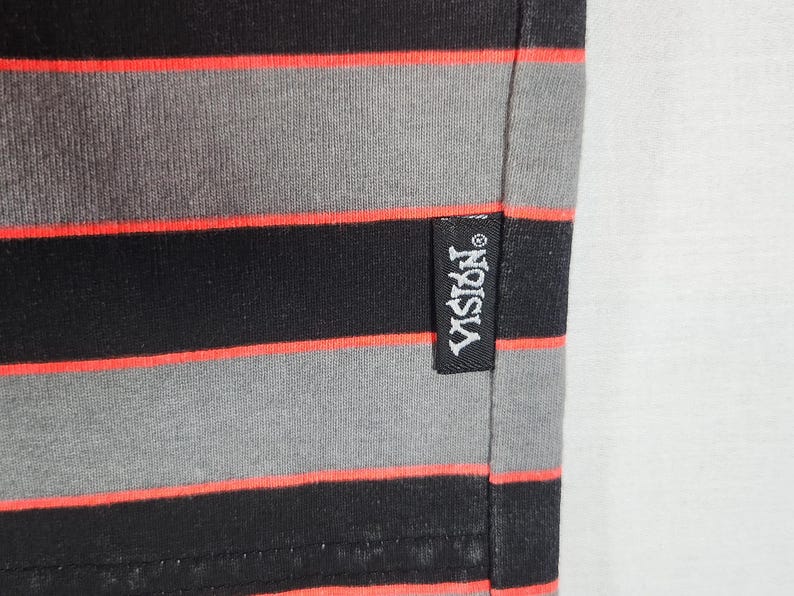 May include: Close-up of a black, gray, and red striped fabric. The fabric has horizontal stripes in black, gray, and red. A black tag with the white text "VISION" is sewn on the fabric.
