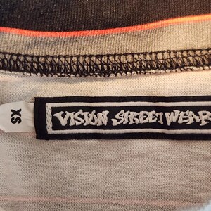 May include: Close-up of a t-shirt tag with the text "VISION STREET WEAR" in white on a black rectangular label. The shirt is a mix of gray, black, and orange. The tag also has an "SX" size label.