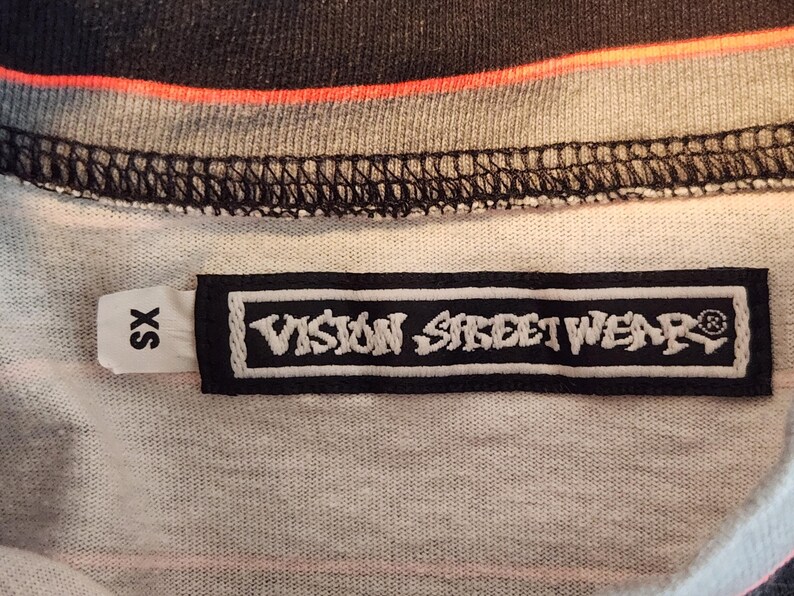 May include: Close-up of a t-shirt tag with the text "VISION STREET WEAR" in white on a black rectangular label. The shirt is a mix of gray, black, and orange. The tag also has an "SX" size label.