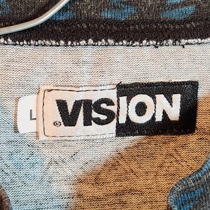 May include: Close-up of a gray and blue patterned fabric with a white label that reads "VISION" in black block letters. The label also has a small "L" on a separate white tag.