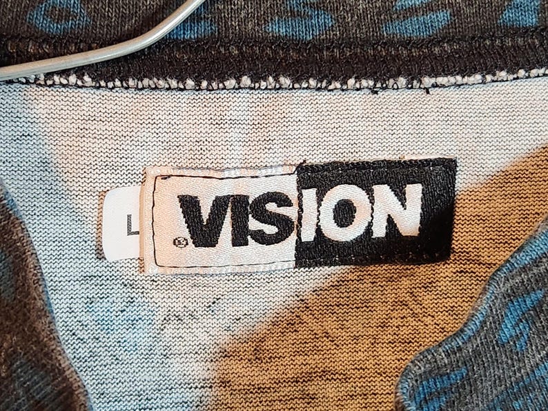 May include: Close-up of a gray and blue patterned fabric with a white label that reads "VISION" in black block letters. The label also has a small "L" on a separate white tag.