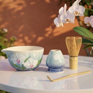 Puede incluir: Un juego de té matcha con un cuenco de cerámica con diseño de orquídeas, un batidor de bambú, un recipiente de té de cerámica y una cuchara de bambú. El cuenco tiene un fondo azul claro con orquídeas blancas y detalles en rosa.