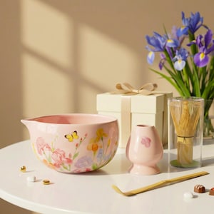 May include: A pink ceramic bowl with a floral design, a small pink ceramic container, a bamboo whisk, and a bamboo scoop. A gift box with a bow and a vase of purple irises are in the background. The bowl has a butterfly and flower design.