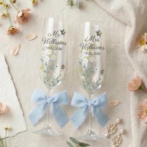 May include: Two champagne flutes, each decorated with floral patterns and the names "Mr. Williams" and "Mrs. Williams" with the date "10.03.2026". Light blue bows adorn the stems. The background features flowers and a cream-coloured cloth.