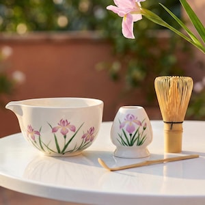 May include: A white ceramic matcha bowl, chawan, and a small ceramic tea cup, chaki, are decorated with purple iris flowers and green stems. A bamboo whisk, chasen, and scoop, chashaku, are also present. The items are on a white table.