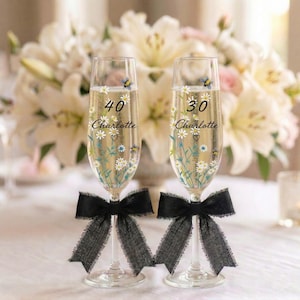 May include: Two champagne flutes with floral designs, the numbers 40 and 30, and the name "Charlotte" are printed on the glasses. Each glass has a black ribbon tied around the base. The background features white flowers.
