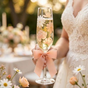 May include: A champagne flute filled with a bubbly beverage, adorned with a peach-colored bow. The glass is decorated with floral designs in shades of peach, white, and yellow. The stemware is held by a person wearing a white lace dress.