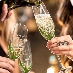 Puede incluir: Copas de champán que se llenan con una bebida espumosa. Las copas están decoradas con un diseño floral y el texto "Melissa Class of 2026". Las copas están siendo sostenidas por personas.
