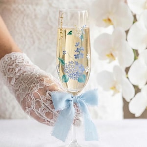 May include: A champagne flute with a light blue bow tied around the stem. The glass is decorated with blue floral designs and the word "Pearles" in gold script. The glass is filled with a sparkling beverage.