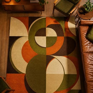 May include: Geometric patterned area rug in shades of orange, brown, green, and beige. The rug features a mid-century modern design with large circular and rectangular shapes. The rug is placed on a wooden floor, with furniture visible in the background.