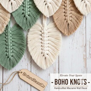May include: Macrame wall decor in the shape of leaves, in shades of cream, beige, and green. The handcrafted pieces are arranged against a white wooden background. A wooden tag with the text "BirdandBeastCo" hangs from one of the leaves. The text "- BOHO KNOTS - Handcrafted Macrame Wall Decor" is also visible.