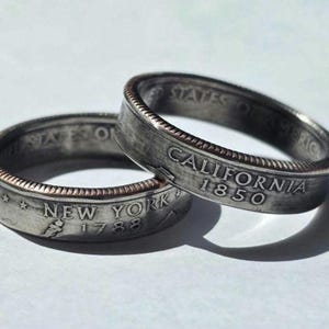 May include: Two silver rings, each crafted from a coin. One ring displays "CALIFORNIA 1850", the other "NEW YORK 1788". The rings have a textured, aged appearance with a contrasting copper-colored edge.