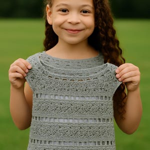 Puede incluir: Un top de ganchillo gris con un dobladillo festoneado y un diseño calado. El top sin mangas tiene un escote redondo y rayas caladas horizontales. El top se muestra en una persona joven sobre un fondo verde borroso.
